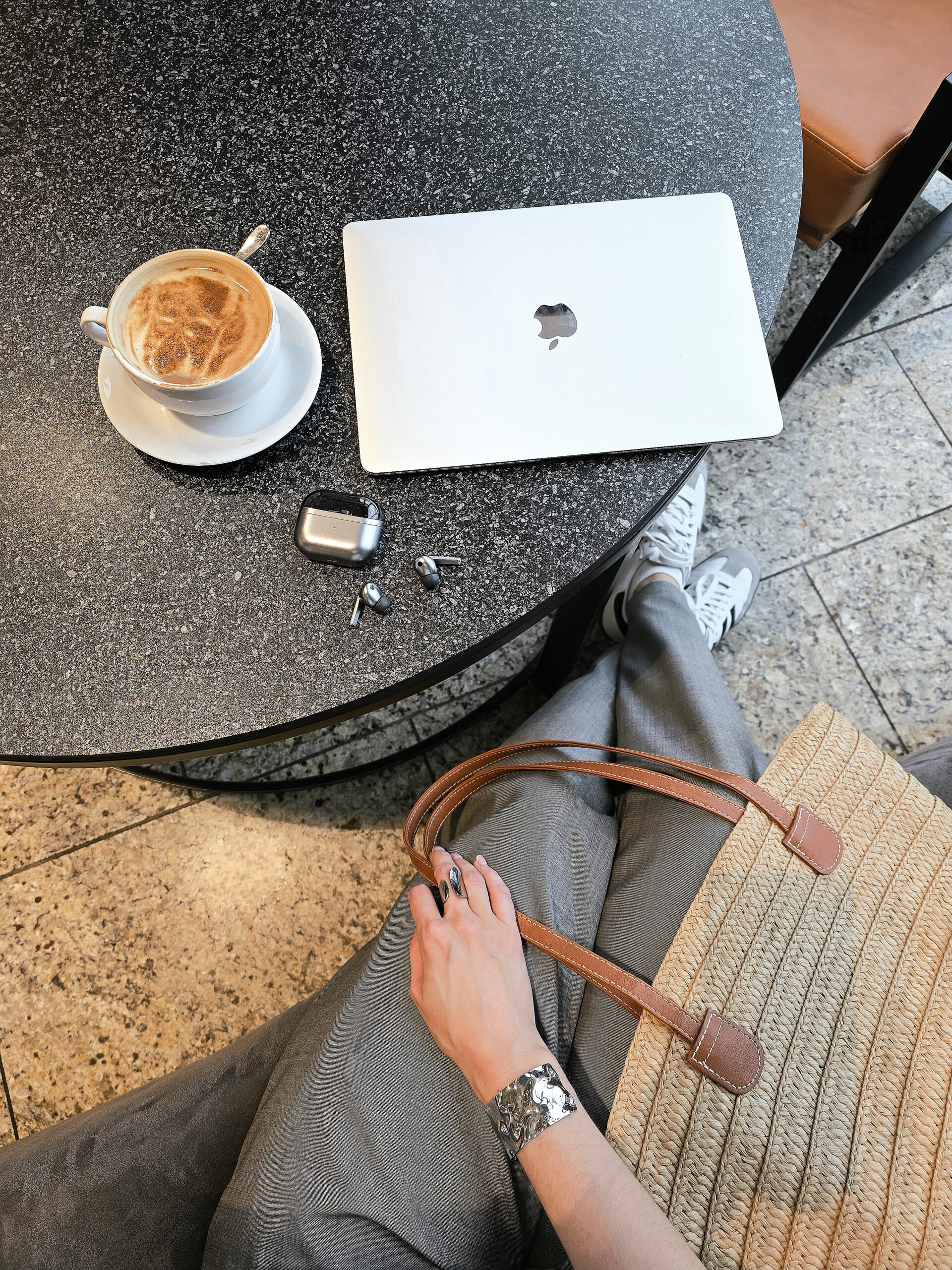 brown bag on a woman's lap, coffee mug with coffee sitting on table beside closed laptop and earbuds